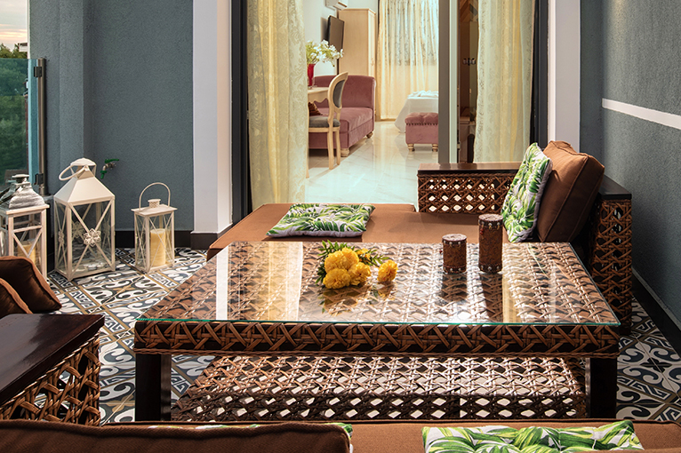 Modern balcony with glass-topped rattan table and comfy corner couch. 
Lanterns and yellow flowers add colour to the setting. 