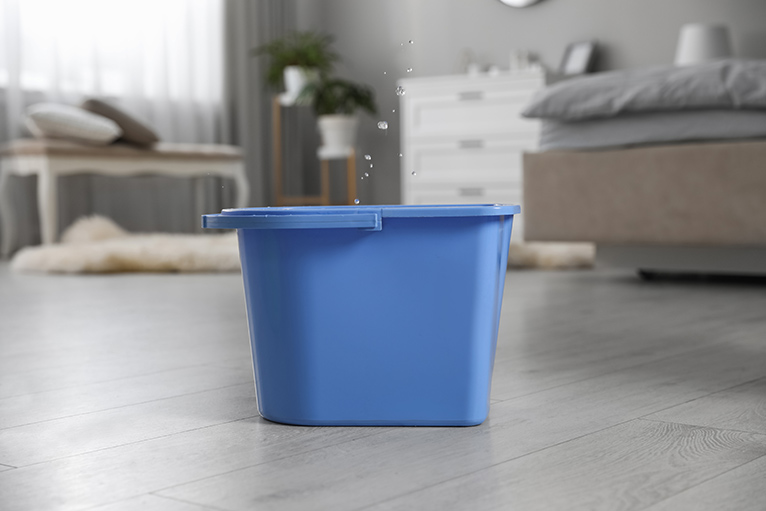 Blue cleaning bucket on floor catching drips from a leaky ceiling. 