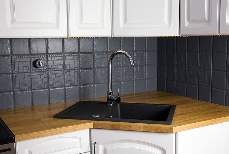 Close up view of new modern kitchen solid wood oak counter with built in black rectangular granite sink, repainted grey colour tile wall. Corner cupboard.